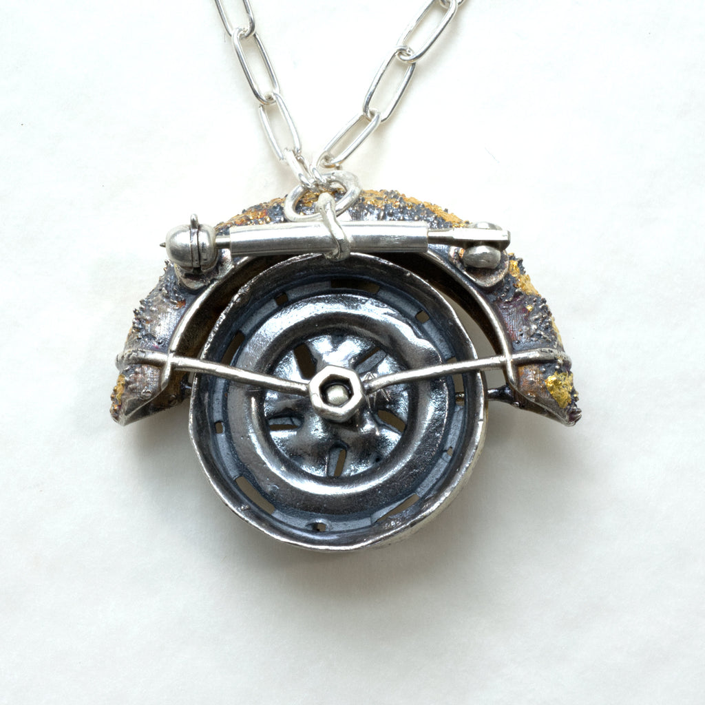 Lux fender brooch pendant with a pink spinel , blackened ssilver. 24k gold fused using keum boo process. The hubcap spins. back with chain