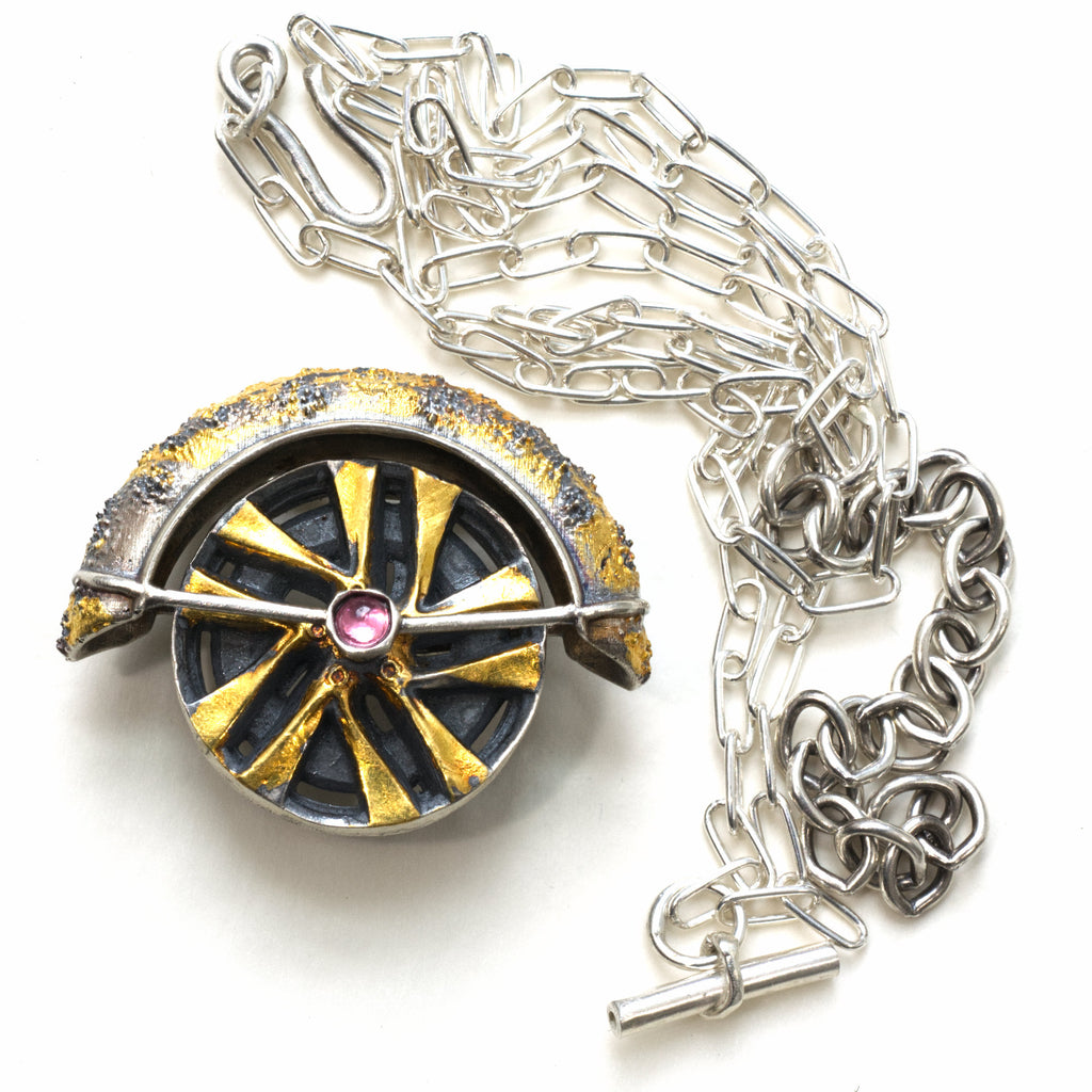 Lux fender brooch pendant with a pink spinel , blackened ssilver. 24k gold fused using keum boo process. The hubcap spins. Frontwith chain at the side.