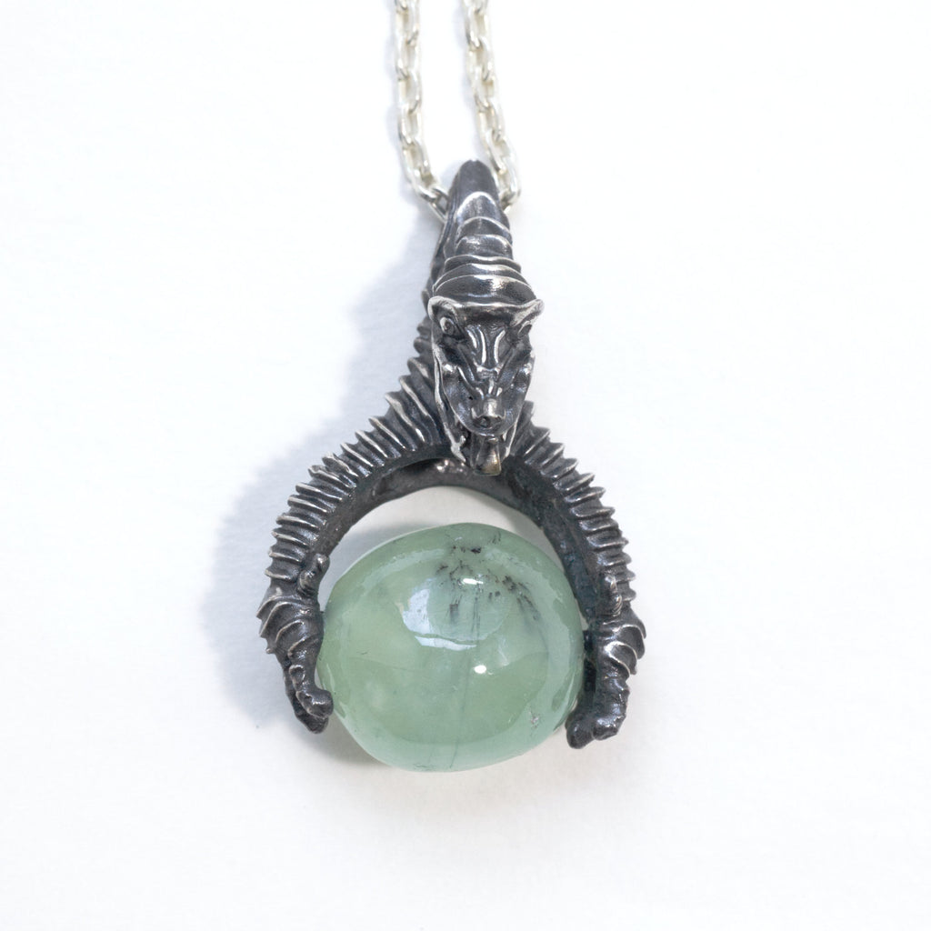 A dragon holding a prehnite sphere like he just caught it. Blackened silver, with a silver chain, front.